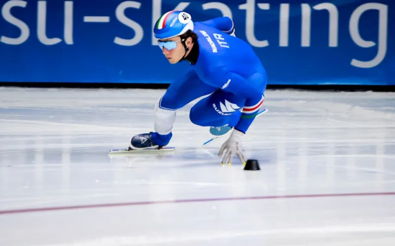Pattinatore in tuta blu si inclina in curva sulla pista di short track.