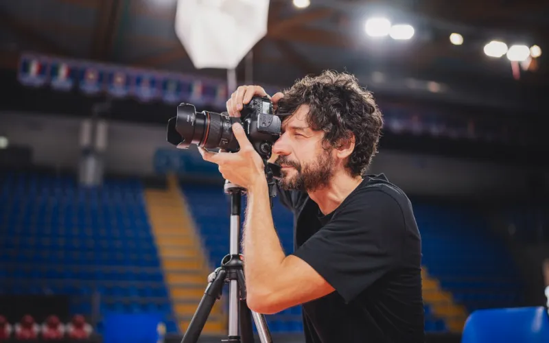Un uomo con una fotocamera al lavoro a bordo campo, davanti alle tribune di un palazzetto.