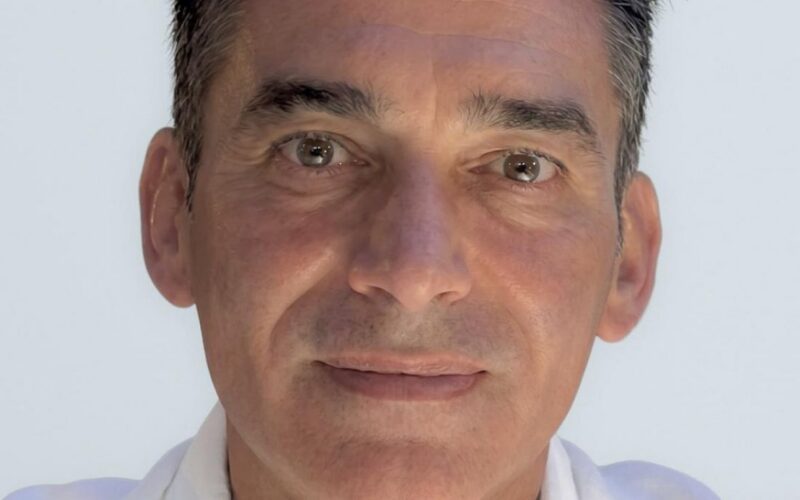 Ritratto frontale di un uomo con camicia bianca su sfondo chiaro.