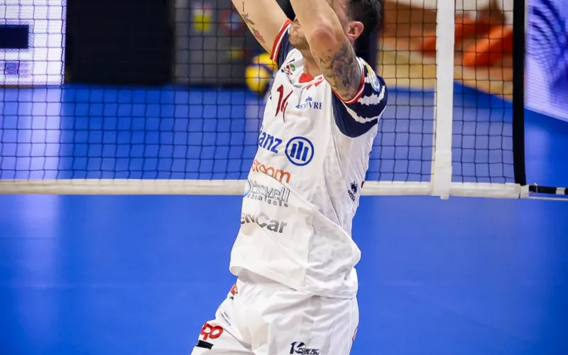 Foto di archivio di una partita di pallavolo, con un giocatore che si prepara a colpire la palla sopra la rete, pubblico e staff visibili sullo sfondo.