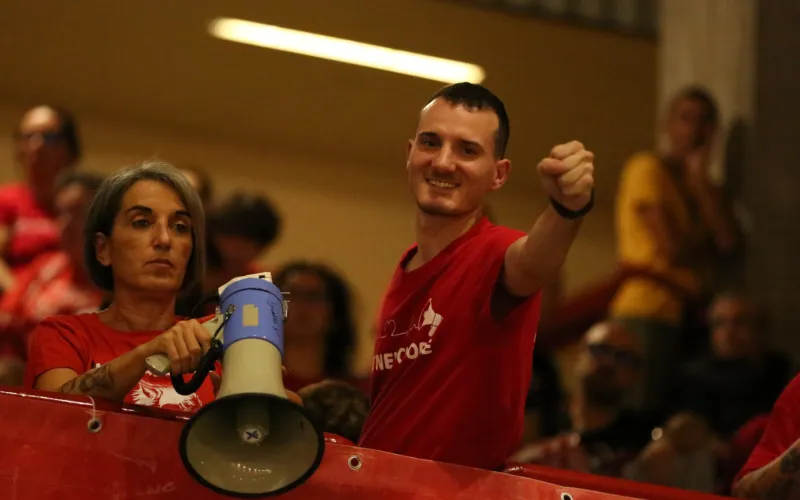 Tifosi durante una partita di pallavolo in palestra, uno con megafono, con magliette rosse.