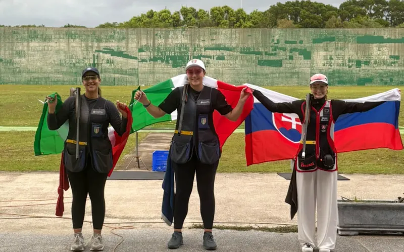 Tre atlete in piedi con bandiere nazionali davanti a una rete di recinzione.