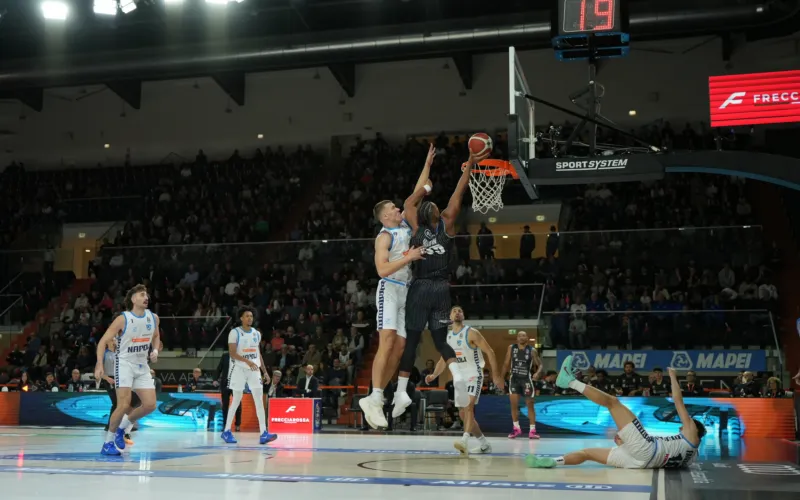 Durante una partita di basket, un giocatore salta vicino al canestro con il pallone; altri giocatori e il pubblico sono visibili sullo sfondo.