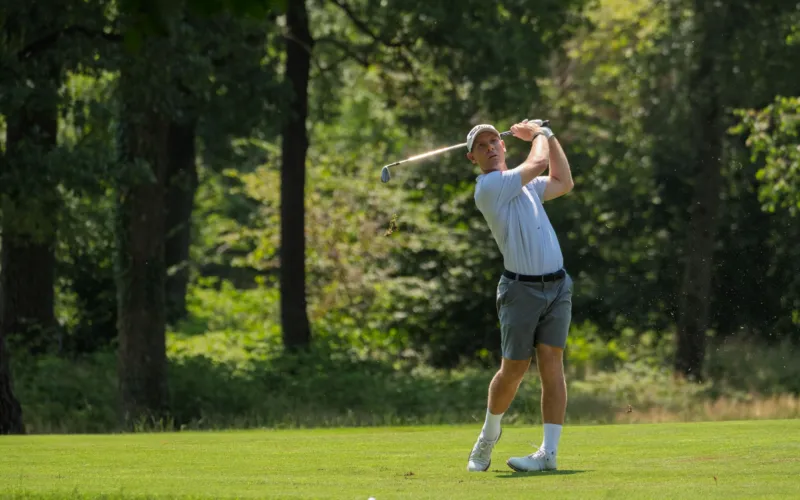 Un golfista completa il movimento del colpo su un campo erboso, con alberi sullo sfondo.