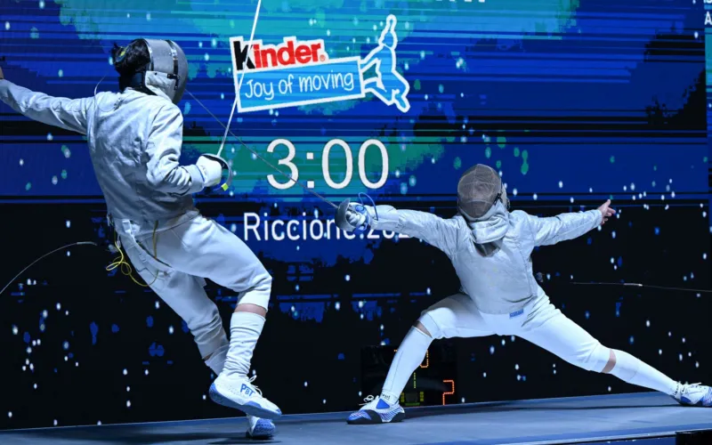 Due schermidori in tenuta bianca si affrontano con le spade su una pedana. Sullo sfondo si leggono “Kinder Joy of Moving” e “Riccione”.